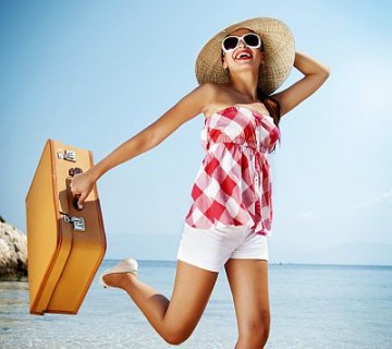 ženska s slamnikom in kovčkom na plaži