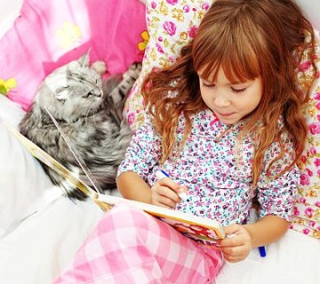 deklica bere knjigo v postelji, poleg leži siva mačka